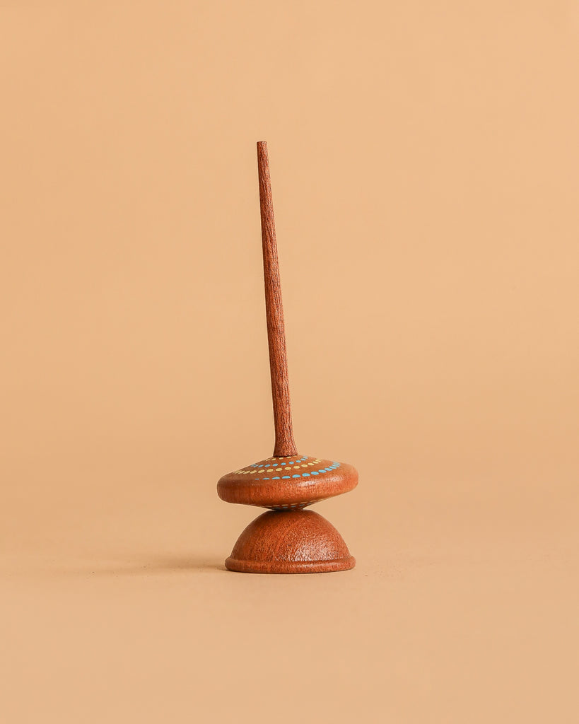 The Wooden Spinning Top - Long Stem, Dots stands upright on a smooth beige surface against a matching beige background.