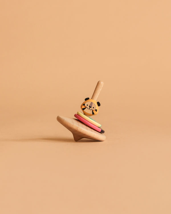 The Wooden Spinning Top - Tiger, made from sustainably sourced wood with colorful rings and a tiger face on top, stands upright on a beige surface against a matching beige background.