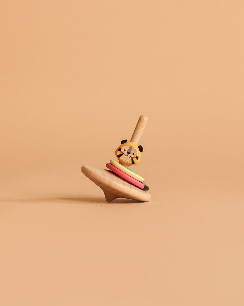 The Wooden Spinning Top - Tiger, made from sustainably sourced wood with colorful rings and a tiger face on top, stands upright on a beige surface against a matching beige background.