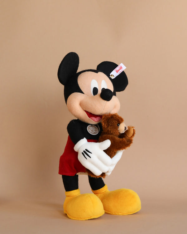 A plush toy of Steiff Collectible Mickey Mouse with Teddy Bear, 12 Inches holding a smaller teddy bear, standing against a plain beige background. Mickey is smiling, wearing his classic red shorts and yellow shoes. This limited edition item celebrates