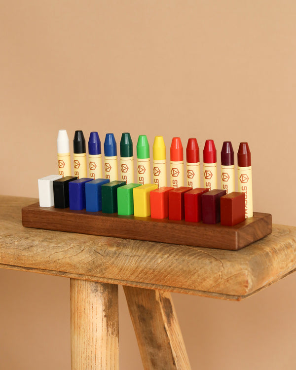 The Stockmar Crayon Set With Wooden Organizer Tray features sixteen vibrant, non-toxic wax crayons in shades like white, black, blue, green, yellow, orange, and red. They are neatly displayed on a wooden stand against a rustic surface and neutral background for all your artistic needs.