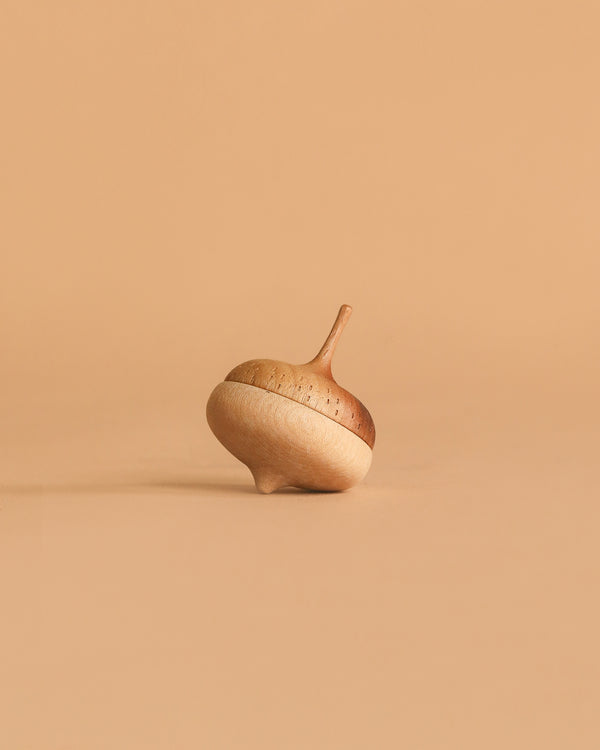 The Wooden Spinning Top - Acorn, crafted from maple walnut and shaped like an acorn, rests on a smooth surface with a solid light brown background—a charming, sustainable toy.