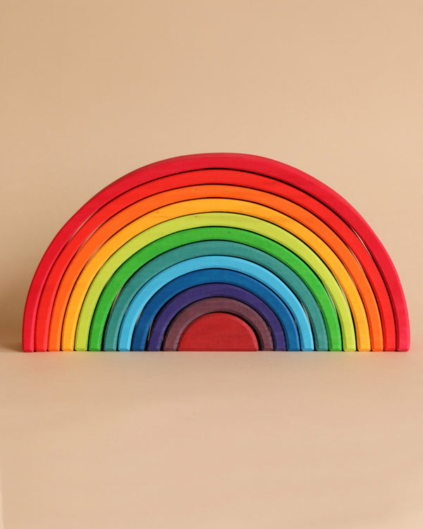 Image of the Grimm's Large Wooden Rainbow, featuring 12 colorful layers in red, orange, yellow, green, blue, and purple. This versatile and beautifully crafted toy is stained with non-toxic water-based colors and arranged in a symmetrical arch against a beige background—an ideal choice for imaginative play.