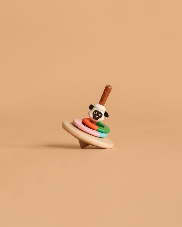 The Wooden Spinning Top - Sheep, crafted from sustainably sourced wood, features colorful rings and a sheep face design on a plain beige background.