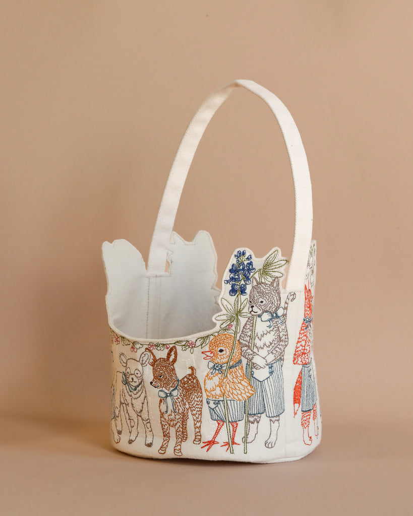 The Coral & Tusk Easter Parade Basket features a charming handle and embroidered animals like sheep, goats, chicks, and rabbits in clothes. This whimsical felt basket is set against a neutral background, adding enchantment to any setting.