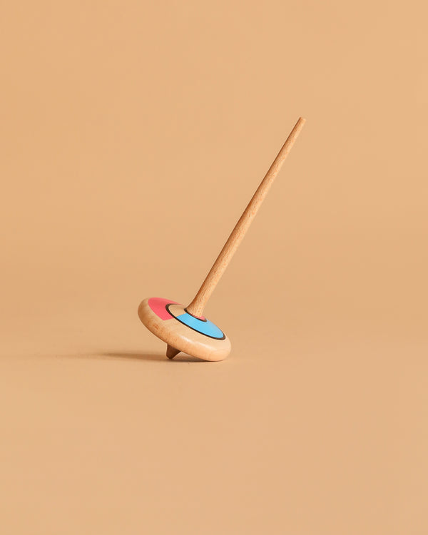 The Wooden Spinning Top - Long Stem, Red & Blue is upright on a light brown surface with a matching beige background, displaying its vibrant circular design.