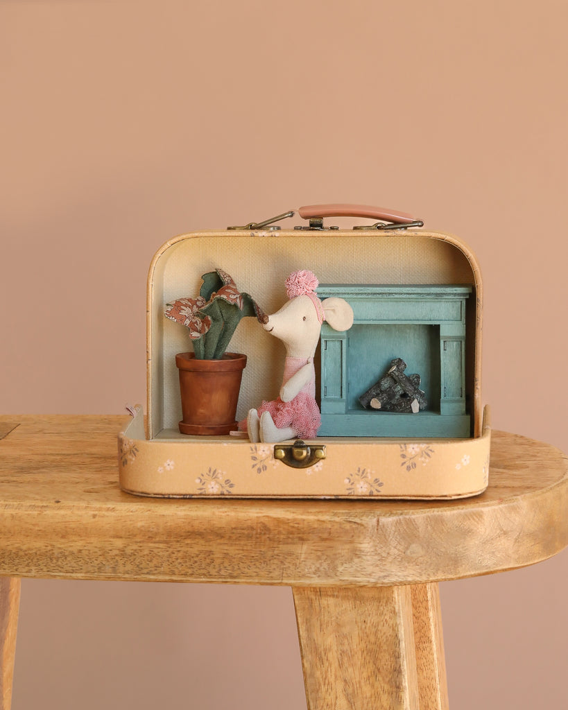 The Maileg Little House Living Room Travel Set rests on a wooden stool, featuring a ballerina mouse in pink, a small flower pot, and a blue fireplace with logs inside an open suitcase. The backdrop is a simple beige wall.