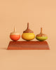 The Wooden Spinning Top Set - Chestnut features three vibrant tops—orange, yellow, and green—standing upright on a smooth wooden stand against a beige background, highlighting this sustainable classic kids' toy.