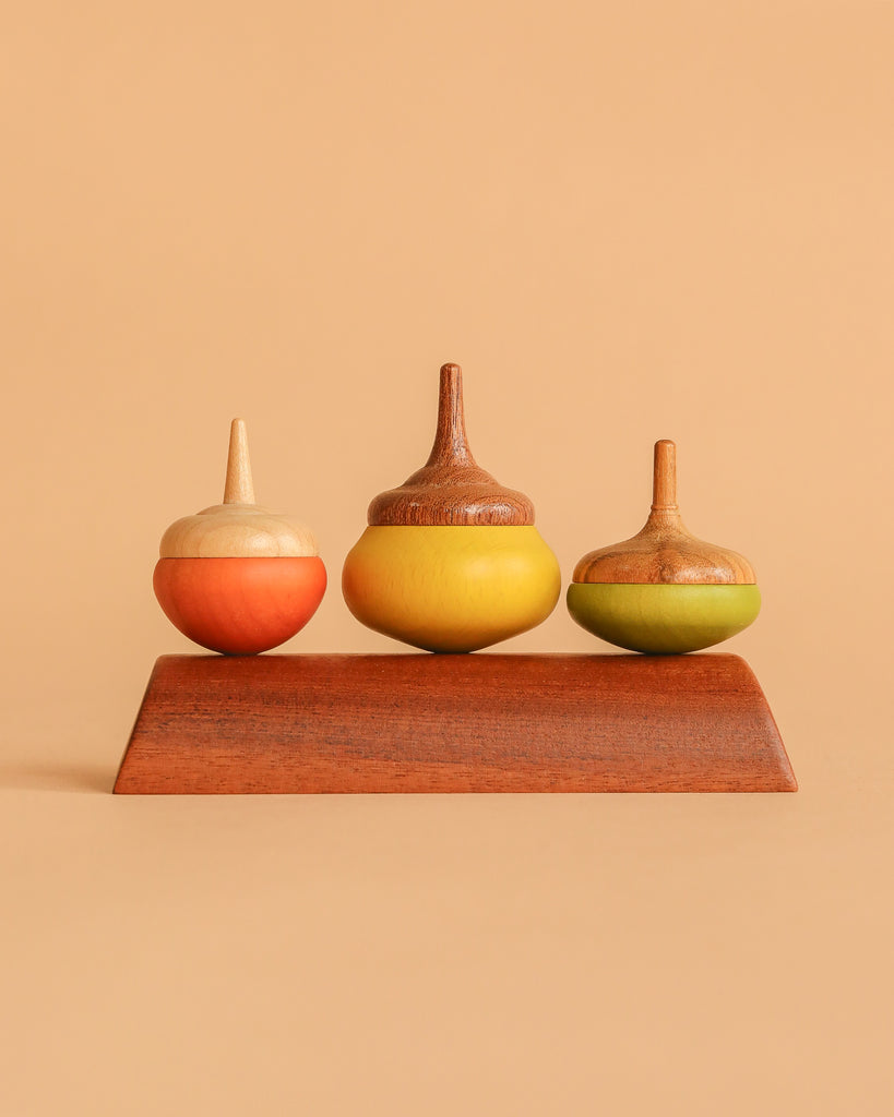 The Wooden Spinning Top Set - Chestnut features three vibrant tops—orange, yellow, and green—standing upright on a smooth wooden stand against a beige background, highlighting this sustainable classic kids' toy.
