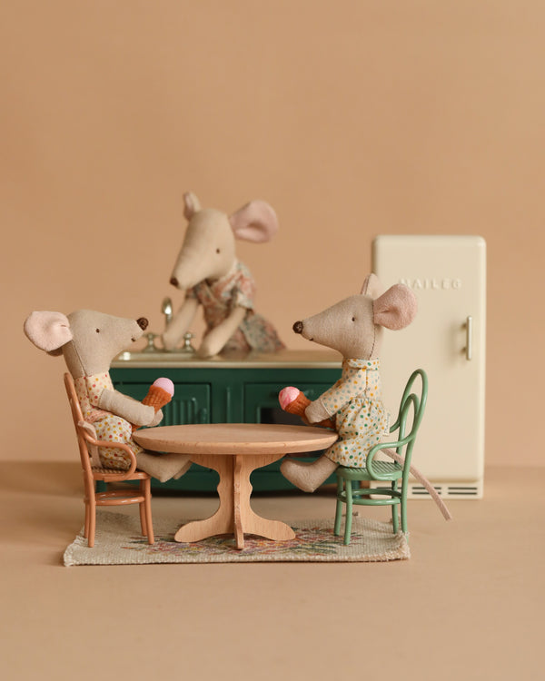Three Maileg mice enjoy their miniature kitchen with the Maileg Kitchen Set: little sister mouse sits at the round table with mugs, another stands behind the counter, while a green cabinet and a white fridge complete the cozy scene.