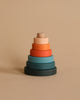 The Mini Wooden Pyramid Stacker - Tropics by SABO Concept is a handmade toy featuring circular rings in various earth-toned colors, arranged in ascending order from largest to smallest against a beige background. Painted with non-toxic paint, the rings showcase shades of dark green, blue, red, orange, and light peach.