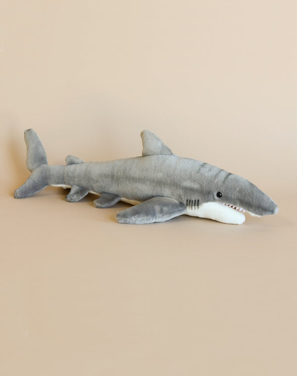 A Great White Shark Stuffed Animal with realistic features and a white underside, resting against a beige background.