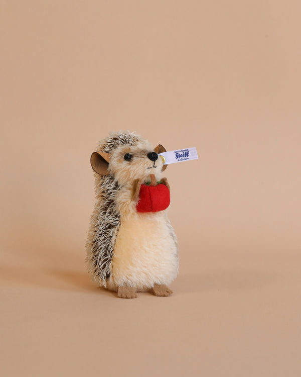 A plush Steiff, Ivo Hedgehog with Felt Apple, 4 inches toy with a fluffy belly and spiky back, holding a red apple and a small white sign with the word "sticker" in blue text, against a soft.