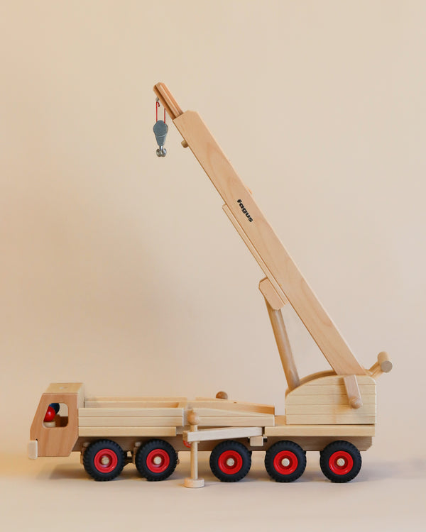 A Fagus Wooden Crane standing on a beige surface with its crane arm extended upwards, featuring multiple red wheels and a small hook at the crane's end.