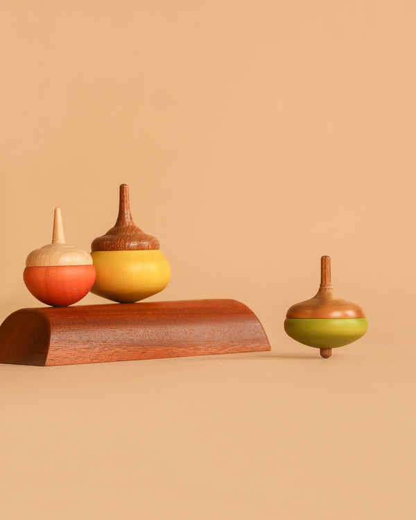 The Wooden Spinning Top Set - Chestnut features three colorful, sustainable wood spinning tops—two on a wooden stand to the left and one spinning on the right—all displayed against a plain beige background.