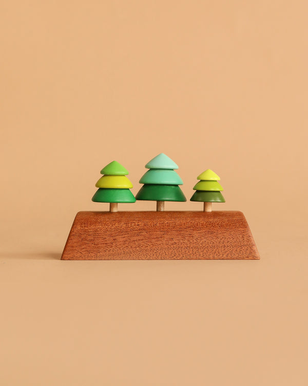 The Wooden Spinning Top Set - Tree features three green wooden trees on a rectangular base, crafted from sustainable wood and designed for timeless play. Set against a light brown background, these colorful toys inspire creativity.