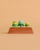 The Wooden Spinning Top Set - Tree features three green wooden trees on a rectangular base, crafted from sustainable wood and designed for timeless play. Set against a light brown background, these colorful toys inspire creativity.