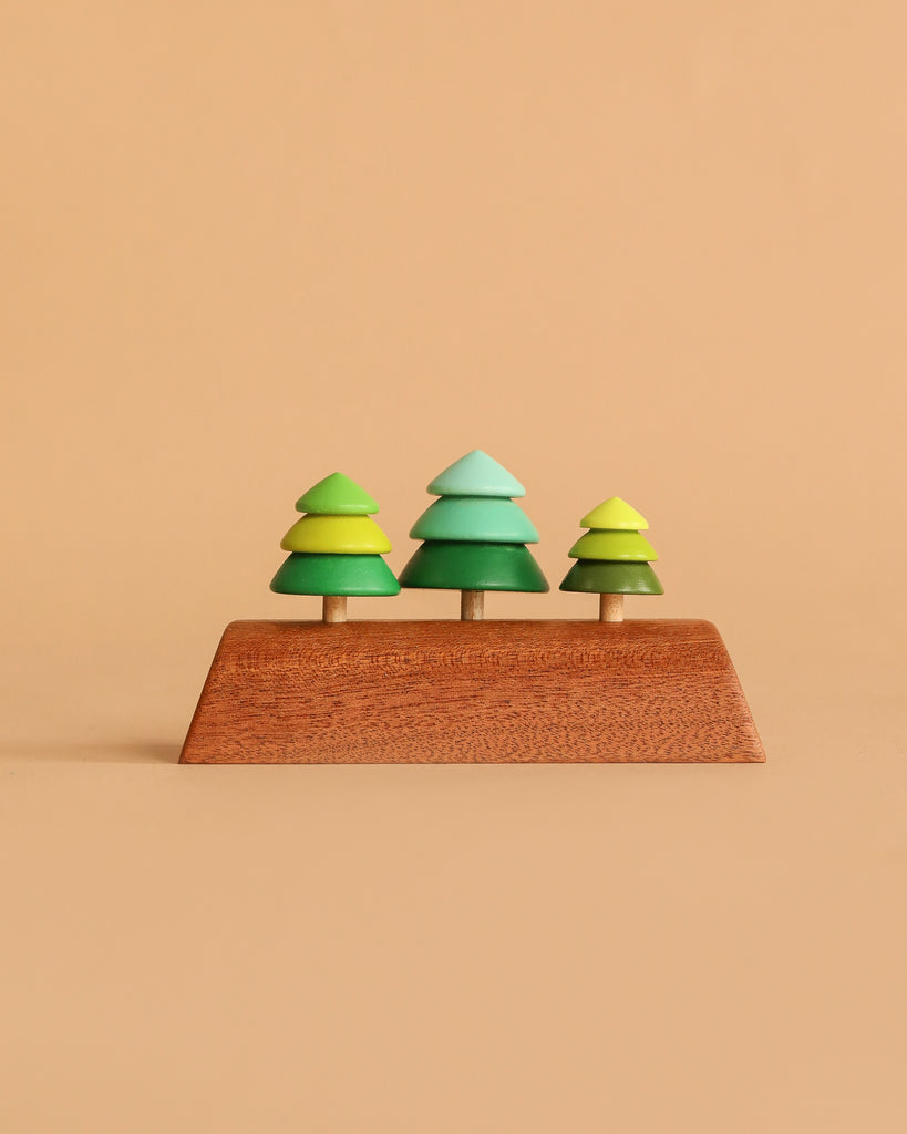 The Wooden Spinning Top Set - Tree features three green wooden trees on a rectangular base, crafted from sustainable wood and designed for timeless play. Set against a light brown background, these colorful toys inspire creativity.