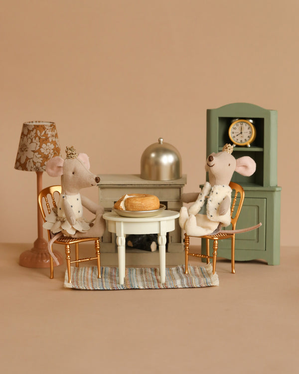 The Maileg Dining Starter Set features two crowned fabric mice at a round table with a cake, tiny chairs, a standing lamp, and a green cabinet with clock. This enchanting dollhouse-like scene contains small parts that may pose choking hazards.