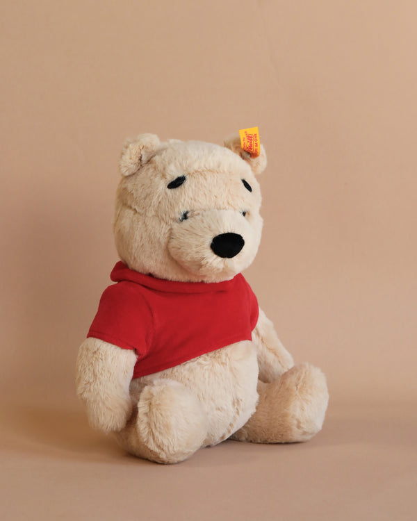 A Steiff, Disney's Winnie the Pooh Jointed Plush, 11 Inches, depicted sitting, wearing his characteristic red shirt and featuring a Button in Ear, against a soft beige background.