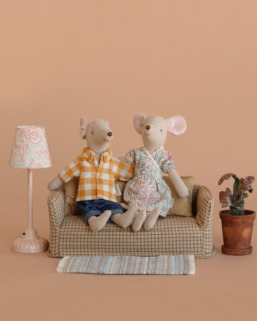 Two Maileg mice from the Maileg Living Room Set sit on a small couch with cute mouse furniture—a vintage floor lamp, potted plant, and rug. One mouse wears a yellow plaid shirt and jeans, the other a floral dress, all set against a tan background.