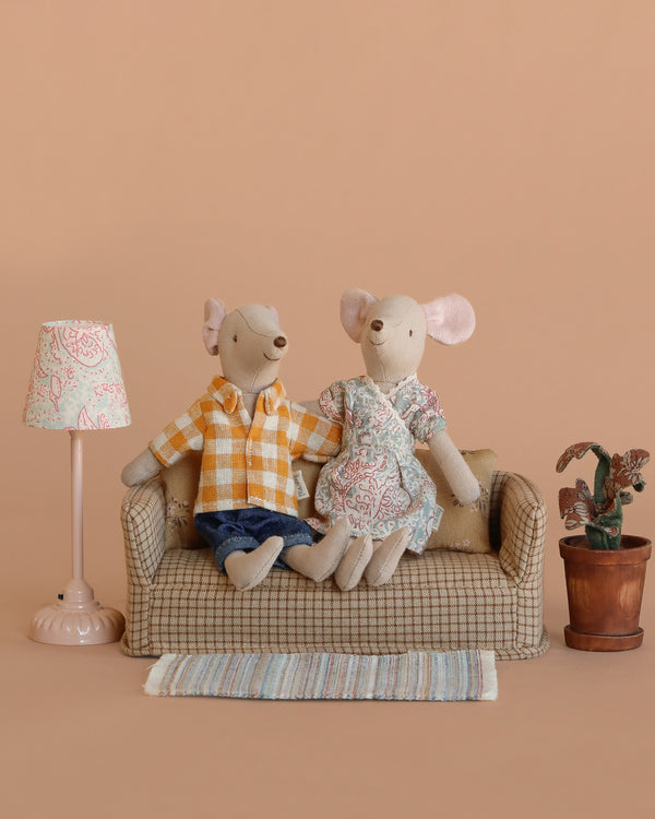 Two Maileg mice from the Maileg Living Room Set sit on a small couch with cute mouse furniture—a vintage floor lamp, potted plant, and rug. One mouse wears a yellow plaid shirt and jeans, the other a floral dress, all set against a tan background.