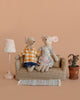 Two Maileg mice from the Maileg Living Room Set sit on a small couch with cute mouse furniture—a vintage floor lamp, potted plant, and rug. One mouse wears a yellow plaid shirt and jeans, the other a floral dress, all set against a tan background.