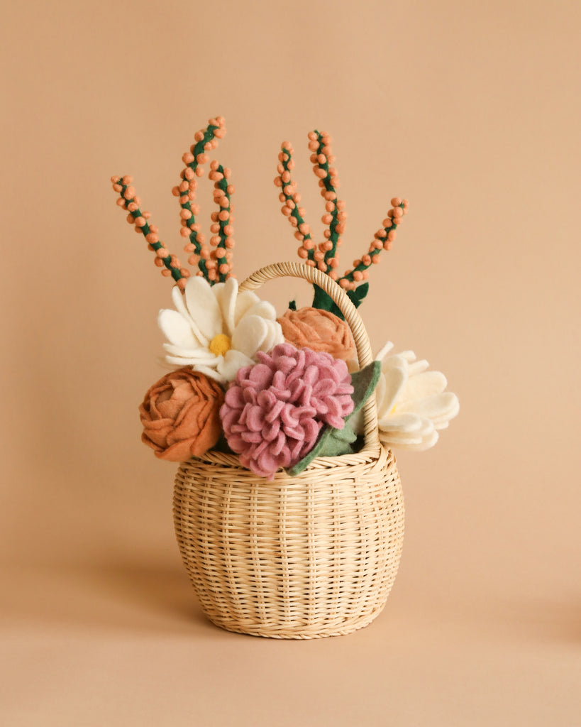 Introducing the Basket of Felt Flowers, a charming small wicker basket brimming with handmade felt flowers in shades of pink, orange, and cream. Enhanced by decorative berry-like branches, this eco-friendly arrangement is crafted using fair wage practices. The warm beige background provides a perfect backdrop to highlight the natural tones of the flowers.