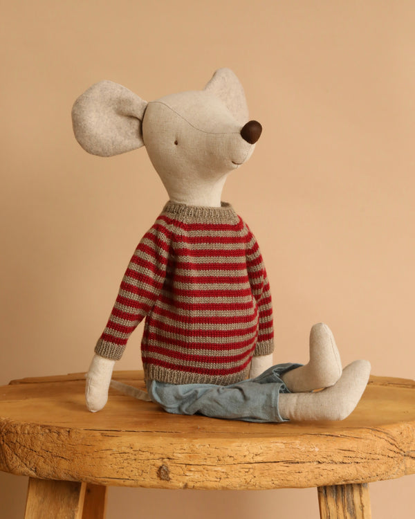 The Maileg Christmas Maxi Mouse, wearing a red and gray sweater and light blue pants, sits on a rustic wooden stool against a plain beige background.