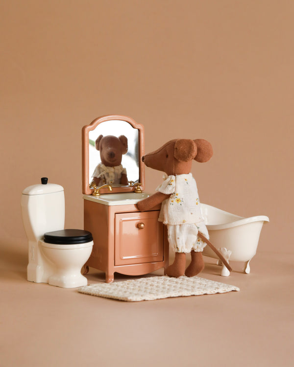 The Maileg Bathroom Set showcases a toy mouse in pajamas standing before a vanity mirror in a delightful miniature bathroom, complete with a white toilet and clawfoot bathtub against a light brown backdrop. Warning: contains small parts; choking hazard for children under 3.