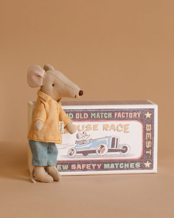 The Maileg "Mouse in a box - Big Brother" is styled in a yellow checkered cotton shirt and denim blue pants, standing beside a vintage-style "House Race" box with a race car illustration, set against a warm, neutral backdrop.