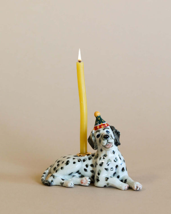 A Dalmatian Cake Topper with a party hat next to a tall, yellow candle, against a soft beige background, showcases heirloom quality craftsmanship.
