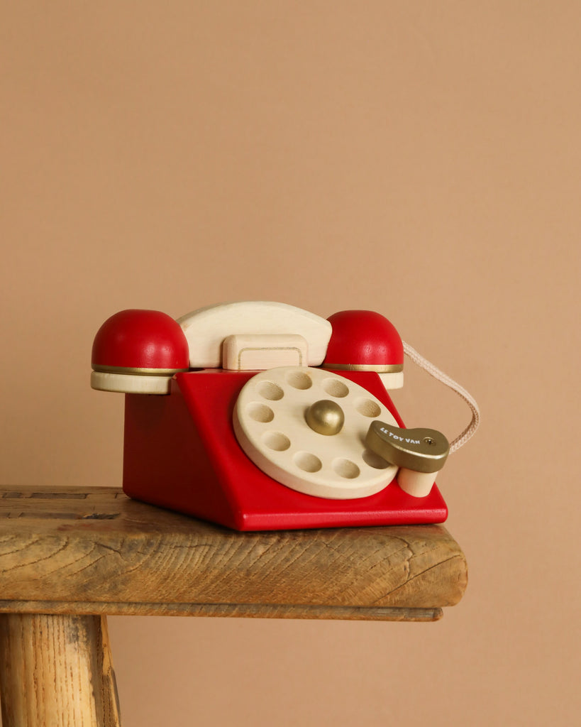 The Vintage Wooden Telephone with a red and cream design and corded receiver rests on a rustic wooden stool against a plain beige background.