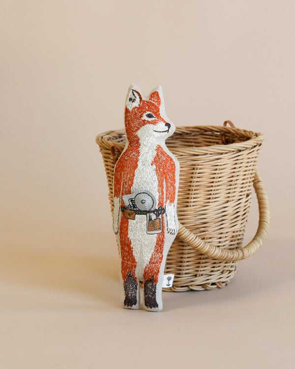 A Coral & Tusk Fox Pocket Doll, painted in white and orange, wearing a belt with strawberries, stands next to a woven basket on a light beige background.
