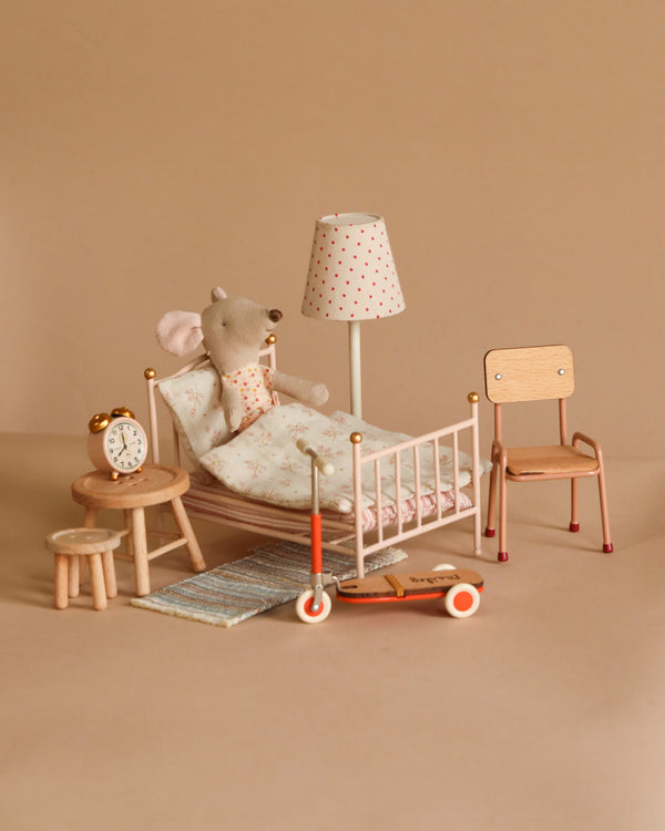 The Maileg Little Sister Bedroom Starter Set features a small plush mouse toy in a bed with floral sheets, complemented by a scooter, clock on a stool, and lamp, all on a beige backdrop for an adorably cozy scene.