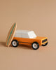 The Candylab Cotswold, a wooden toy vehicle with a minimalist design, is painted in orange and white and positioned against a beige background. Leaning against the car is a small, wooden surfboard featuring a green stripe along its length, adding a playful, beach-themed element to the scene.