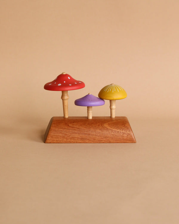 Three Wooden Mushroom Spinning Tops of varying sizes and colors (red, purple, and yellow), crafted from responsibly-sourced wood, mounted on a rectangular wooden base, set against a plain beige background.