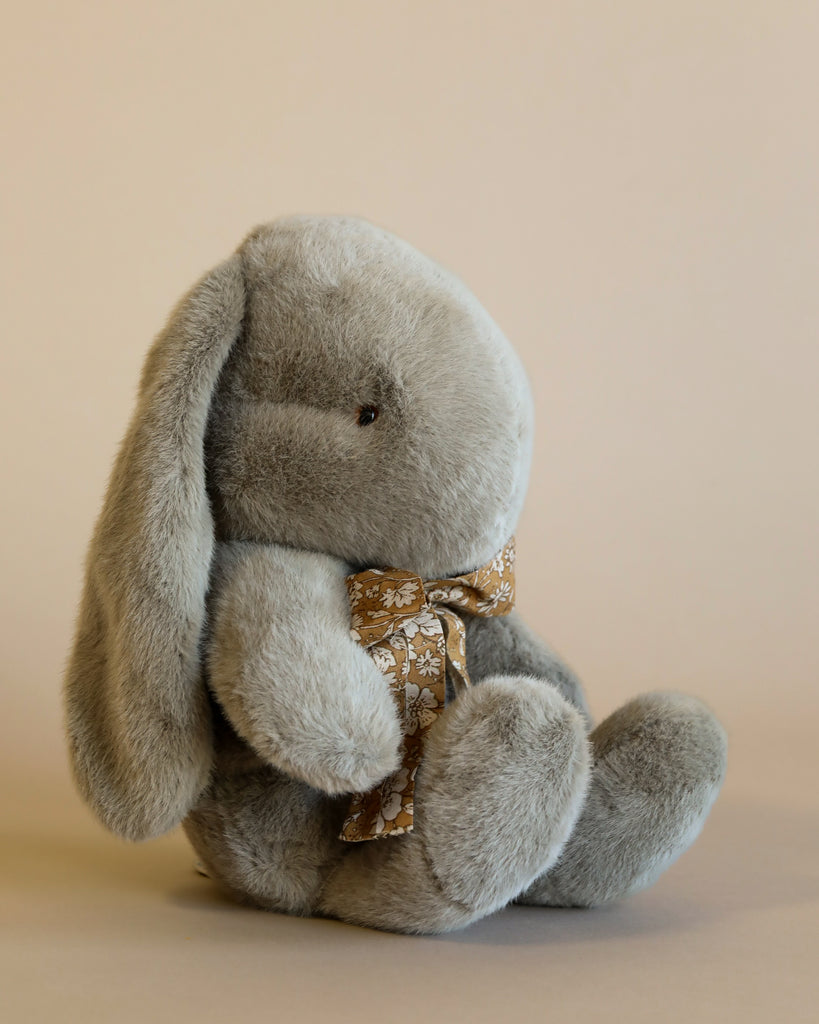 A Maileg Medium Plush Bunny with light gray fur is sitting upright against a plain beige background. The bunny, made from soft plush fabric, has long floppy ears and wears a beautiful bow decorated with a white floral pattern around its neck.