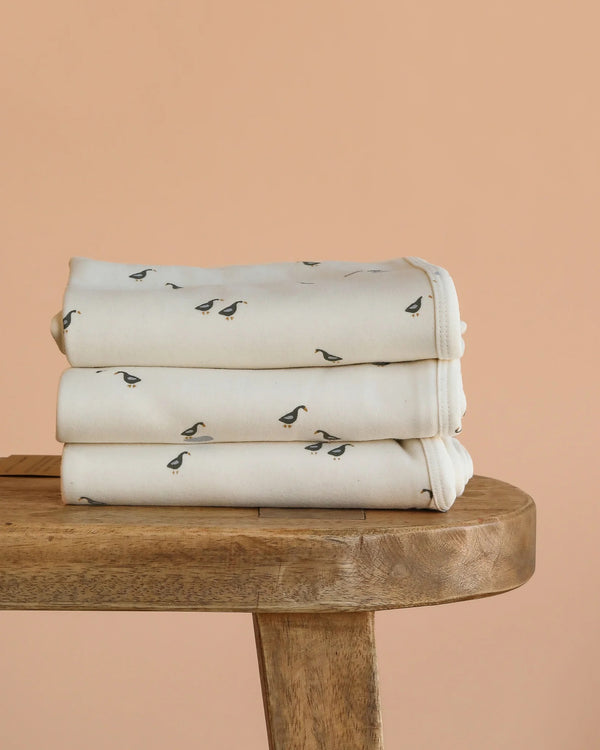 Three cream-colored Organic Baby Blanket – Runner Ducks, featuring small black duck prints, are neatly folded and stacked on a wooden table against a plain peach background.