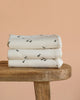 Three cream-colored Organic Baby Blanket – Runner Ducks, featuring small black duck prints, are neatly folded and stacked on a wooden table against a plain peach background.