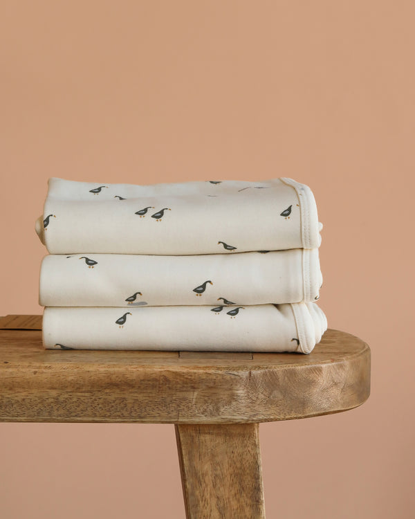 Three neatly folded Organic Baby Blanket – Runner Ducks, featuring a charming black duck pattern on cream-colored organic cotton, are stacked on a wooden bench against a plain beige background.