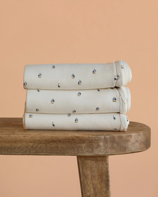 Three folded Organic Baby Blanket – Schooner, featuring small sailboat patterns and made from GOTS-certified organic cotton, are stacked on a wooden bench against a peach-colored background.