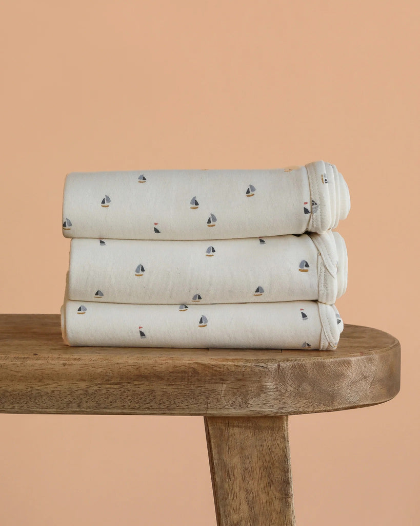 Three folded Organic Baby Blanket – Schooner, featuring small sailboat patterns and made from GOTS-certified organic cotton, are stacked on a wooden bench against a peach-colored background.