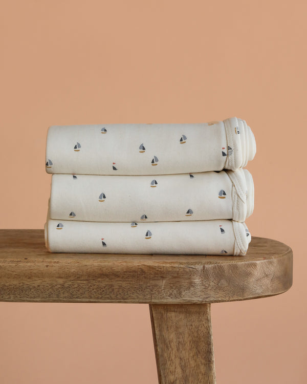 Three neatly folded Organic Baby Blanket – Schooner, featuring small sailboat patterns and made from GOTS-certified organic cotton, are stacked on a wooden bench against a peach-colored background.