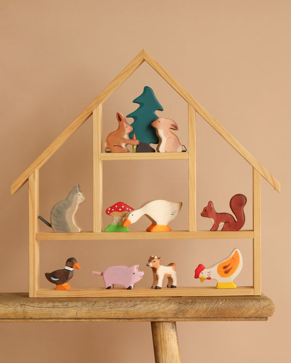 A Holztiger Handmade Wooden Animal Play Set—including a rabbit, squirrel, cat, duck, goose, pig, cow, hen, and tree—adds European charm to a wooden dollhouse shelf set against a beige background.