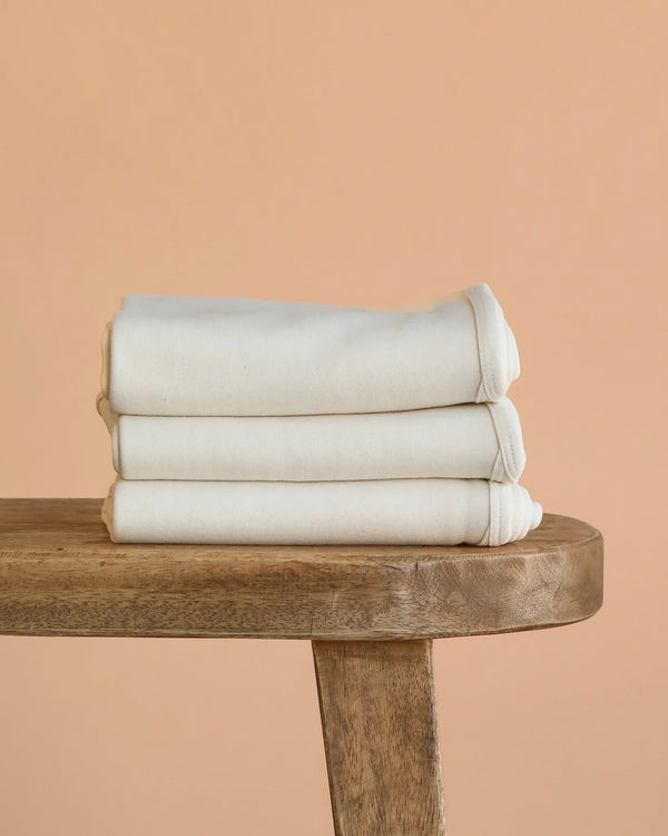 An Organic Baby Blanket – Natural, GOTS-certified and neatly folded, rests on a wooden bench against a peach-colored background.