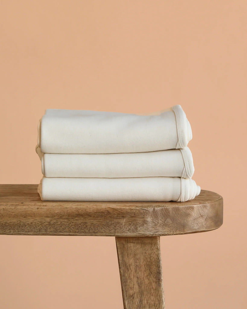 An Organic Baby Blanket – Natural, GOTS-certified and neatly folded, rests on a wooden bench against a peach-colored background.