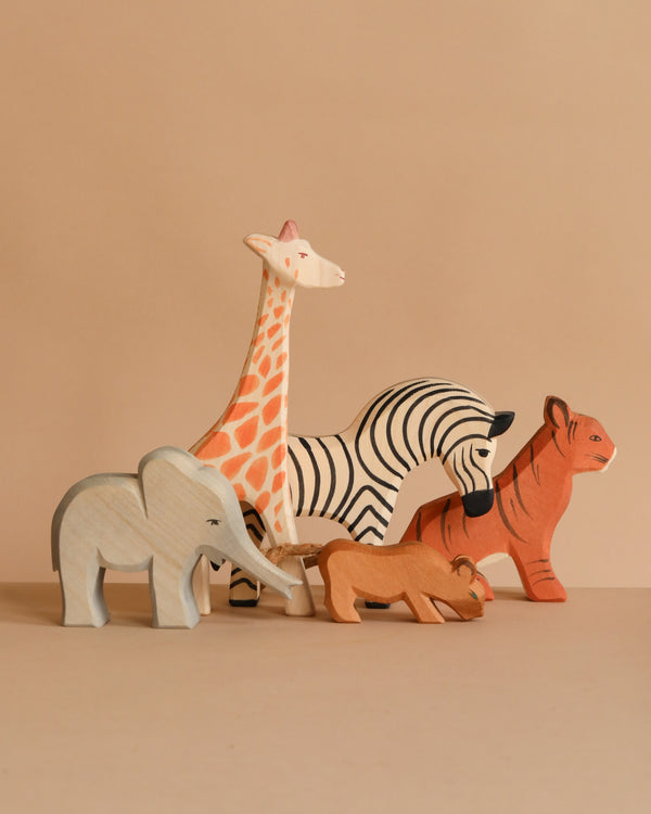 The Ostheimer Handmade Wooden Safari Animals Set features five non-toxic, handcrafted wooden figures—a giraffe, zebra, elephant, lion, and tiger—painted in natural colors and arranged together on a neutral surface with a beige background.