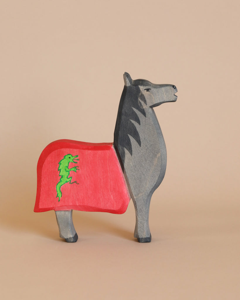 An Ostheimer Horse for Black Knight, painted in shades of gray, stands against a beige background. The wooden toy horse is draped in a red blanket adorned with a green dragon emblem. Its head is slightly turned to the side, showing a stoic expression, perfect for imaginative play and handcrafted wooden toys enthusiasts.