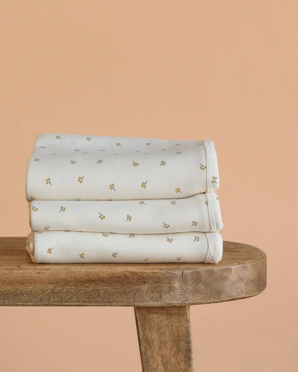 A neatly folded stack of Organic Baby Blanket – Gold Sprig, made from GOTS certified organic cotton and featuring small yellow and green floral patterns, sits on a wooden bench against a peach background.